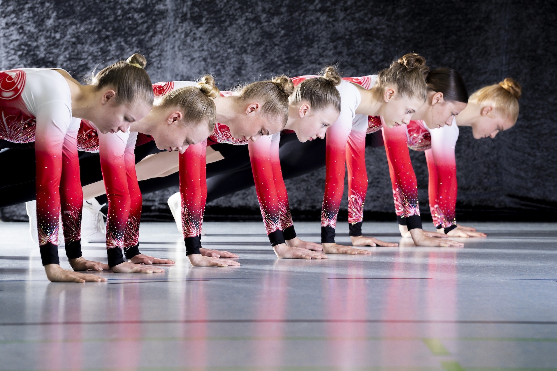 Sechs Gymnastinnen in &auml;hnlicher Sportbekleidung beugen sich beim Training, H&auml;nde auf dem Boden, in einer synchronen Haltung.