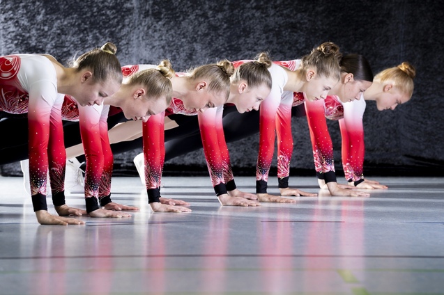 Sechs Gymnastinnen in &auml;hnlicher Sportbekleidung beugen sich beim Training, H&auml;nde auf dem Boden, in einer synchronen Haltung.