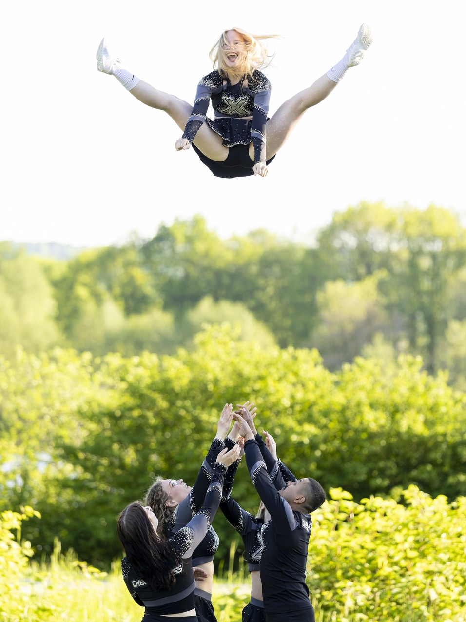 Cheerleader springt in den Luft, umringt von Teamkollegen, die sie mit gestreckten Armen fangen. Gr&uuml;nes Umfeld im Hintergrund.