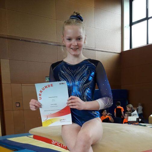 Mädchen in blauer Gymnastikanzug sitzt auf einem Sprungtisch und hält eine Urkunde in der Hand.