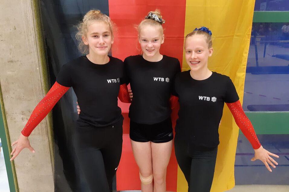 Drei Turnerinnen in schwarzen T-Shirts mit &bdquo;WTB&ldquo; posieren vor einer Deutschlandflagge.