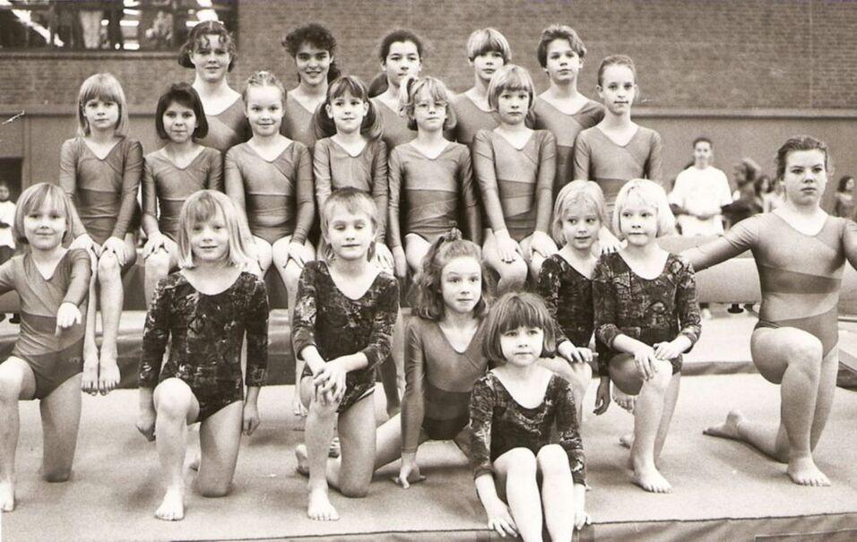 Gruppe von jungen Turnerinnen in Ballettanzanzügen, posierend auf einer Gymnastikmatte in einem Sportzentrum.