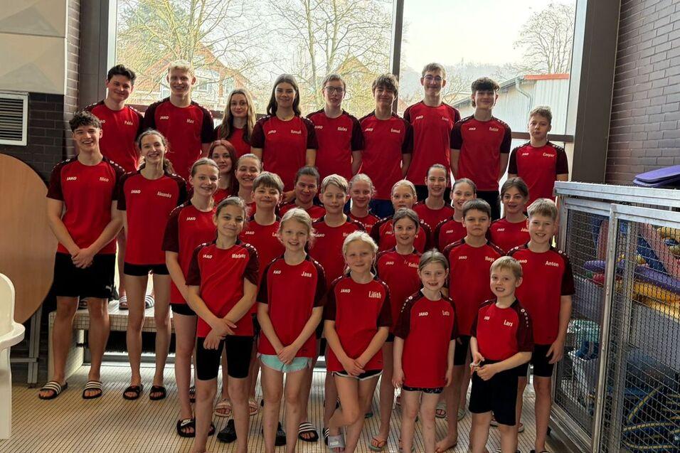 Gruppenfoto von Schwimmern in roten Trikots, aufgereiht vor großen Fenstern in einem Hallenbad.