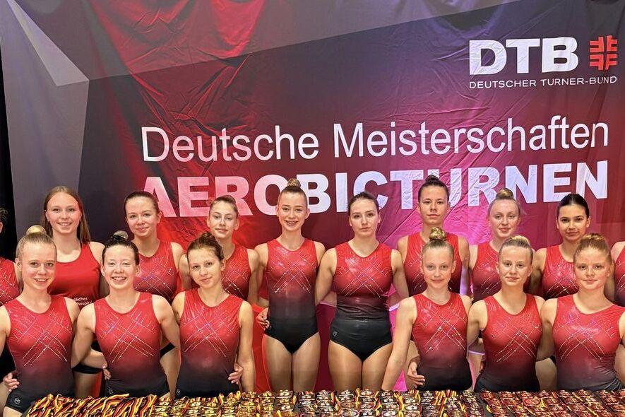 Gruppe von Sportlerinnen in roten Trikots vor einem Banner der Deutschen Meisterschaften im Aerobicturnen.