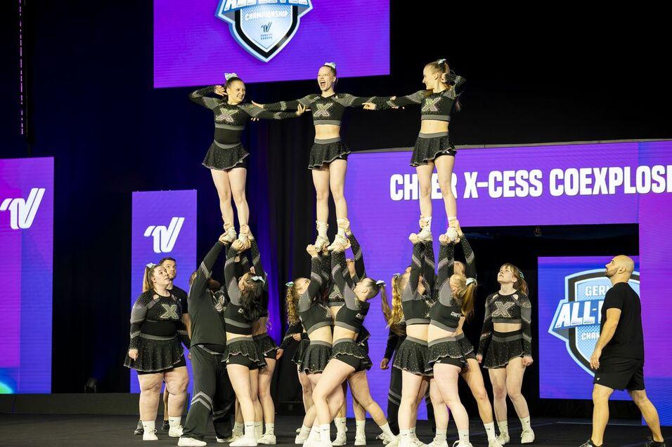Cheerleading-Team führt eine Stunt-Formation mit mehreren Mitgliedern auf einer Bühne mit beleuchteten Hintergrundanzeigen aus.