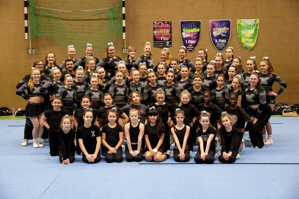 Gruppenfoto von Cheerleadern in schwarzen Uniformen, auf einem blauen Boden, umgeben von Pokalen und Bannern.