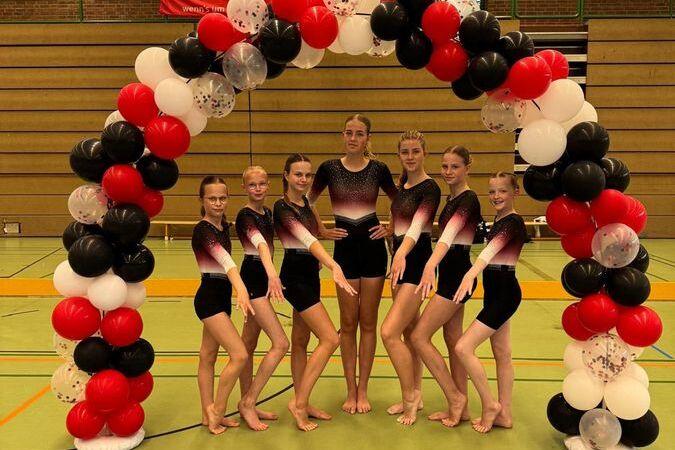 Gruppe junger Turnerinnen in Schwarz-Rot vor einem bunten Luftballonbogen in einer Sporthalle.