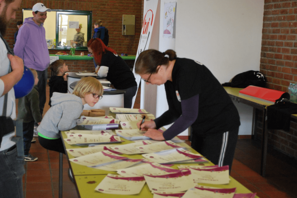 Frau mit Stift schreibt an einem Tisch, während ein Kind zuschaut; um sie liegen zahlreiche Urkunden.