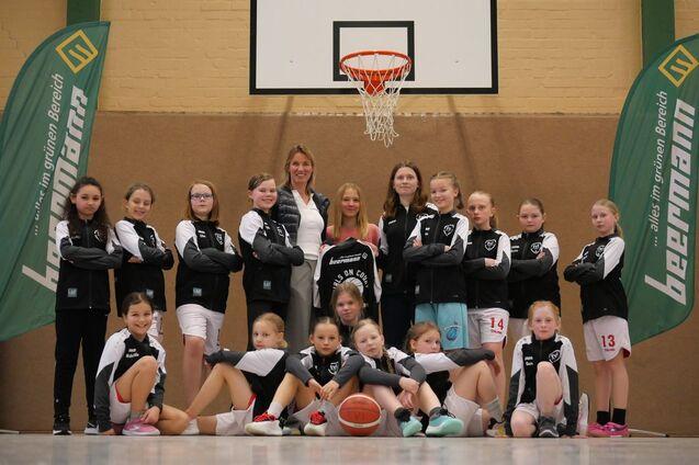 Gruppenfoto eines Basketballteams mit Trainerinnen, in Sportkleidung, vor einem Basketballkorb in einer Halle.