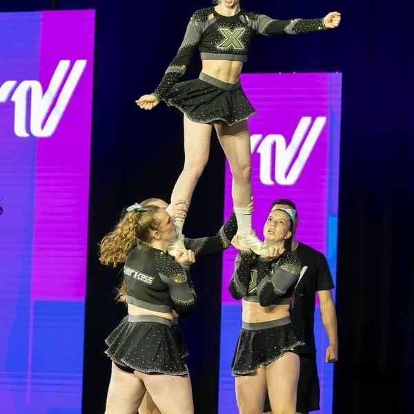 Cheerleaderin wird von zwei Teamkolleginnen in die Luft gehalten, während eine weitere Person im Hintergrund zuschaut.