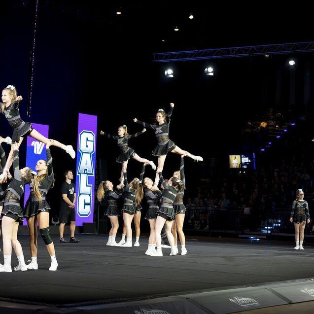 Gruppenchoreografie mit Cheerleadern in schwarz, die Akrobatik und Stunts auf einer Bühne ausführen.