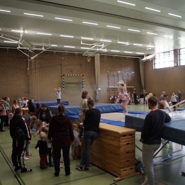 Gruppe von Kindern und Erwachsenen in einer Turnhalle, verschiedene Aktivitäten an Sprungtischen und Matten.
