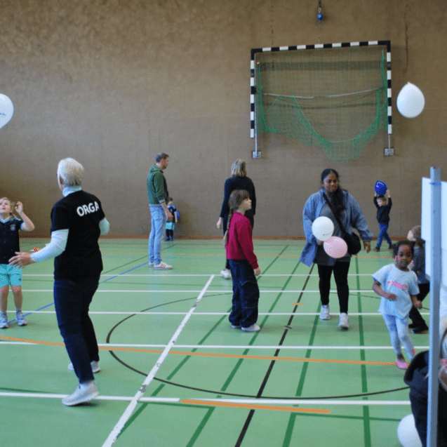 Gruppenspiele in einer Sporthalle mit Kindern, Erwachsenen und Luftballons auf einem grünen Sportboden.
