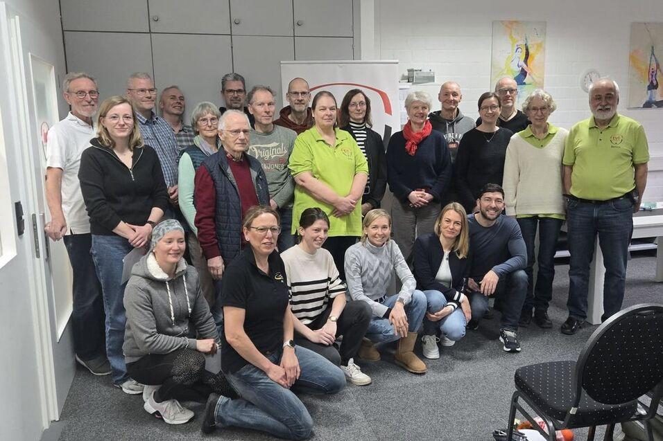 Gruppenfoto von 24 Personen in einem Raum, die lächeln, mit einem Banner im Hintergrund und Stühlen im Vordergrund.