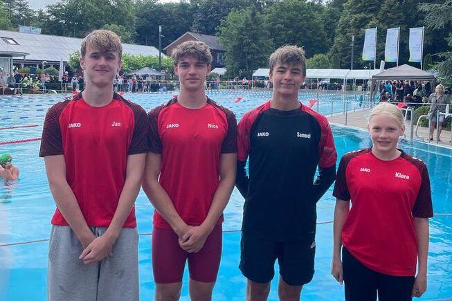 Vier junge Schwimmer in roten Sportoutfits stehen am Beckenrand eines Freibades, im Hintergrund sind Schwimmer und Zuschauer sichtbar.
