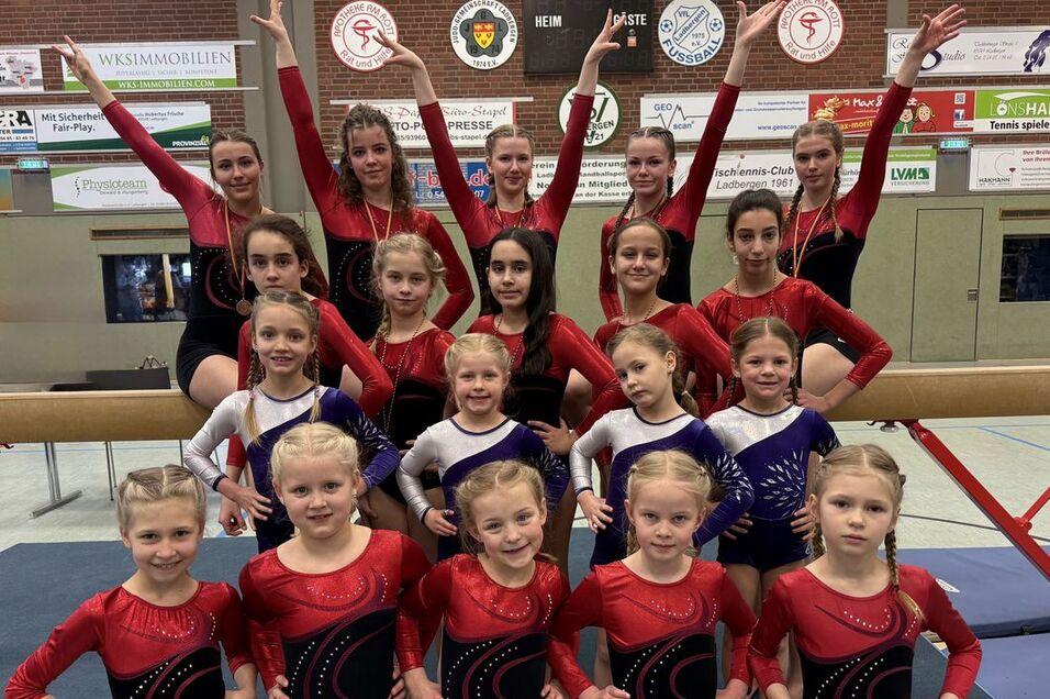 Gruppe von Turnerinnen in Gymnastikanzügen posiert auf einer Turnhalle, einige stehen hinter, andere sitzen vorne.