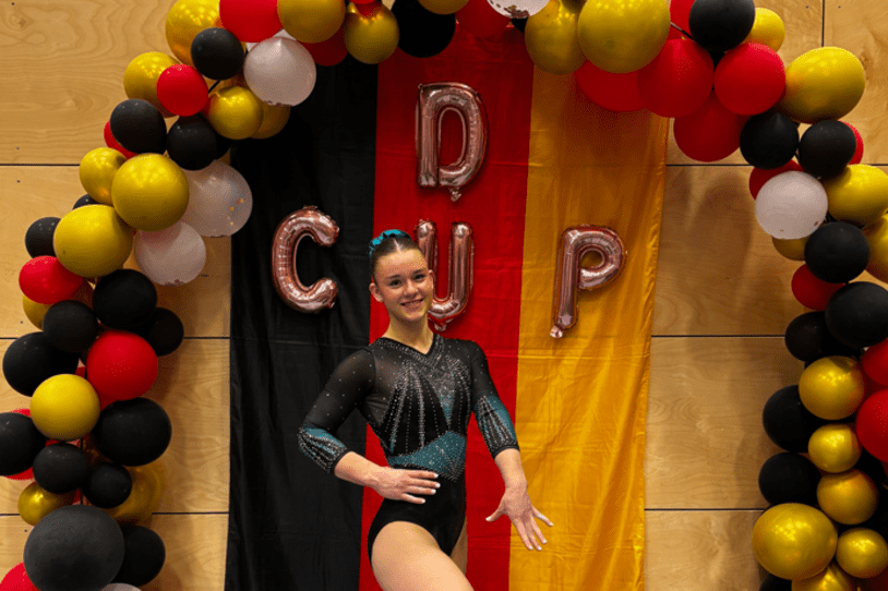 Sportlerin in eleganter Gymnastikanzug posiert vor rot-schwarz-goldenem Hintergrund mit Luftballons und "CUP"-Buchstaben.