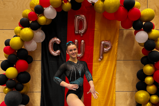 Sportlerin in eleganter Gymnastikanzug posiert vor rot-schwarz-goldenem Hintergrund mit Luftballons und "CUP"-Buchstaben.