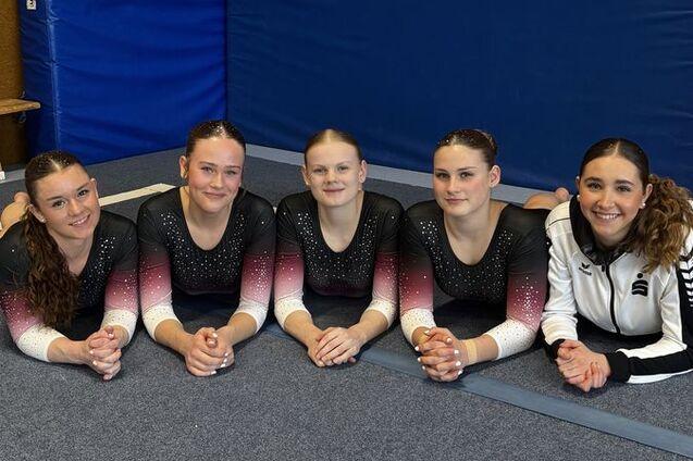 Fünf Mädchen in Sportoutfits liegen auf einer Gymnastikmatte in einer Turnhalle und posieren.