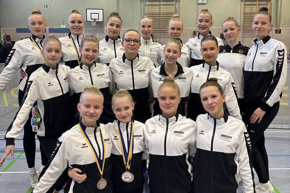 Gruppenshot von 15 Gymnastinnen in schwarzen und weißen Trainingsanzügen mit Medaillen, in einer Halle stehend und kniend.