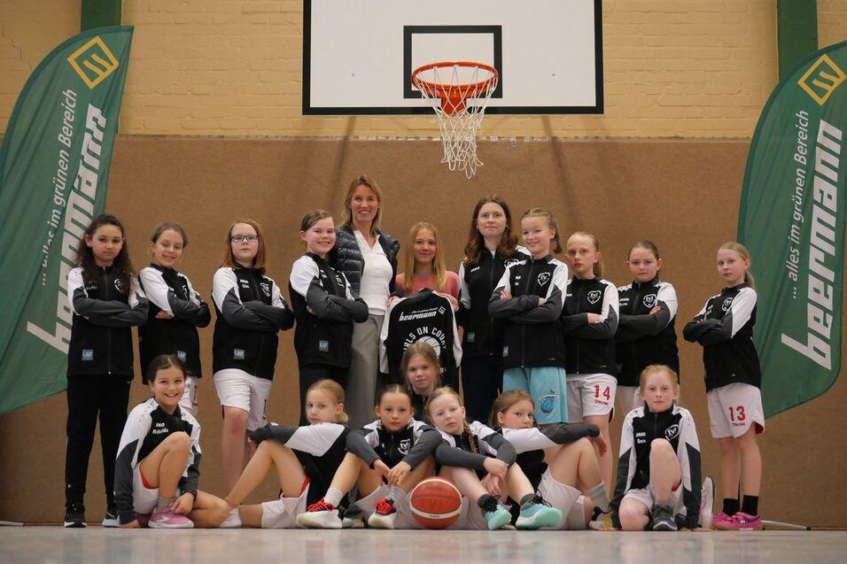Gruppenfoto eines Basketballteams mit Trainerinnen, in Sportkleidung, vor einem Basketballkorb in einer Halle.