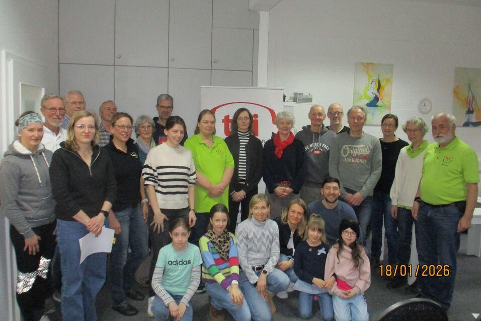 Gruppenfoto von 29 Personen, die in einem Raum posieren, einige tragen grüne Shirts, mit einem Banner im Hintergrund.