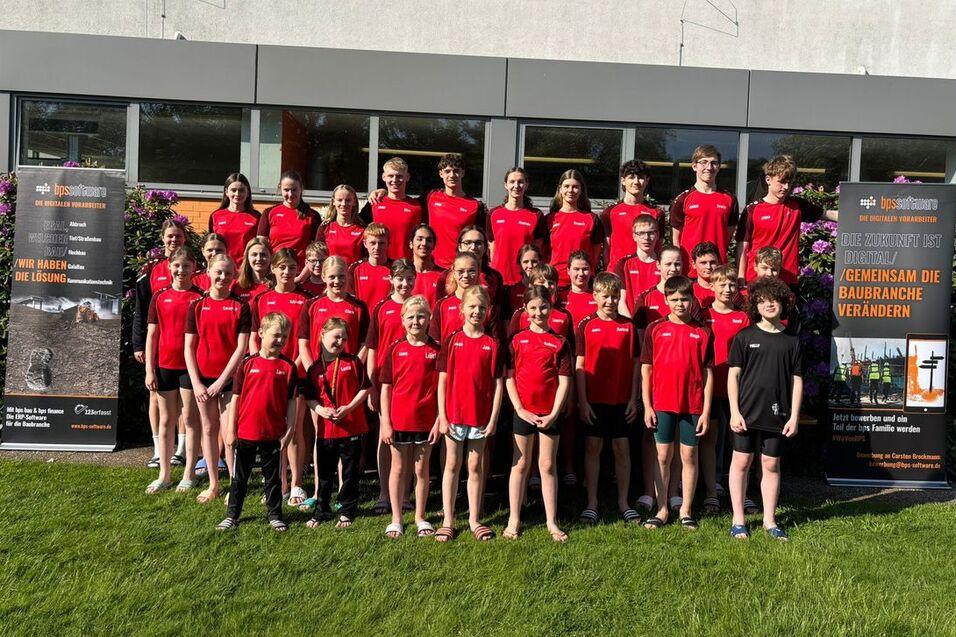 Gruppe von Sportlern in roten Shirts steht vor einem Gebäude mit Bannern, im Freien auf grünem Rasen.
