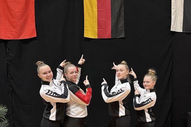 Fünf Sportlerinnen in einheitlicher Kleidung posieren vor einem schwarzen Hintergrund mit mehreren Flaggen, darunter die deutsche.