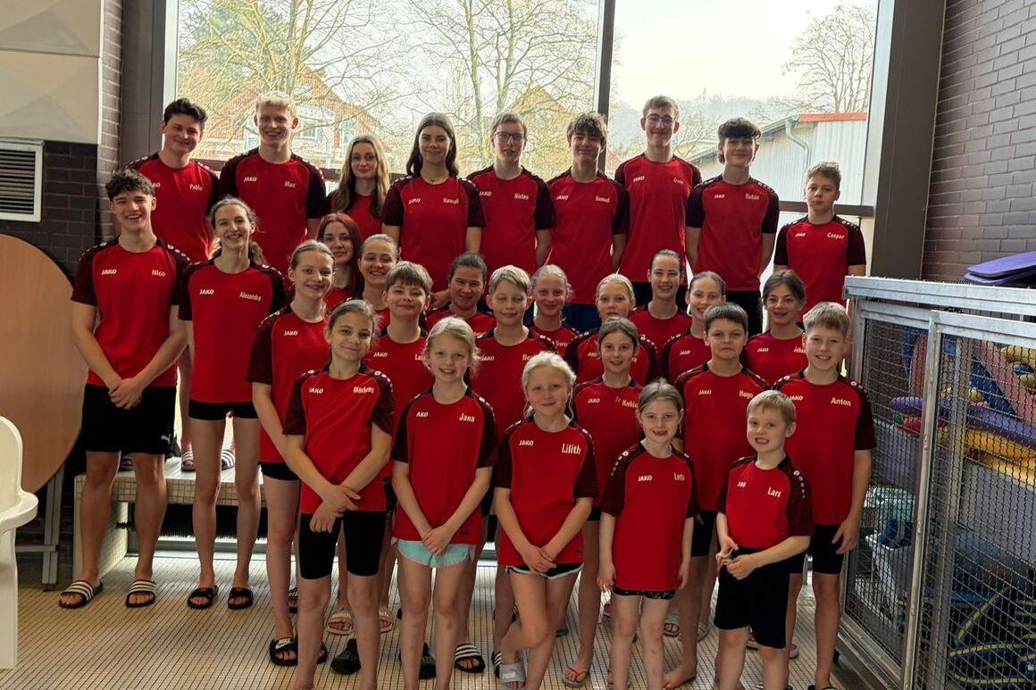 Gruppenfoto von Schwimmern in roten Trikots, aufgereiht vor großen Fenstern in einem Hallenbad.