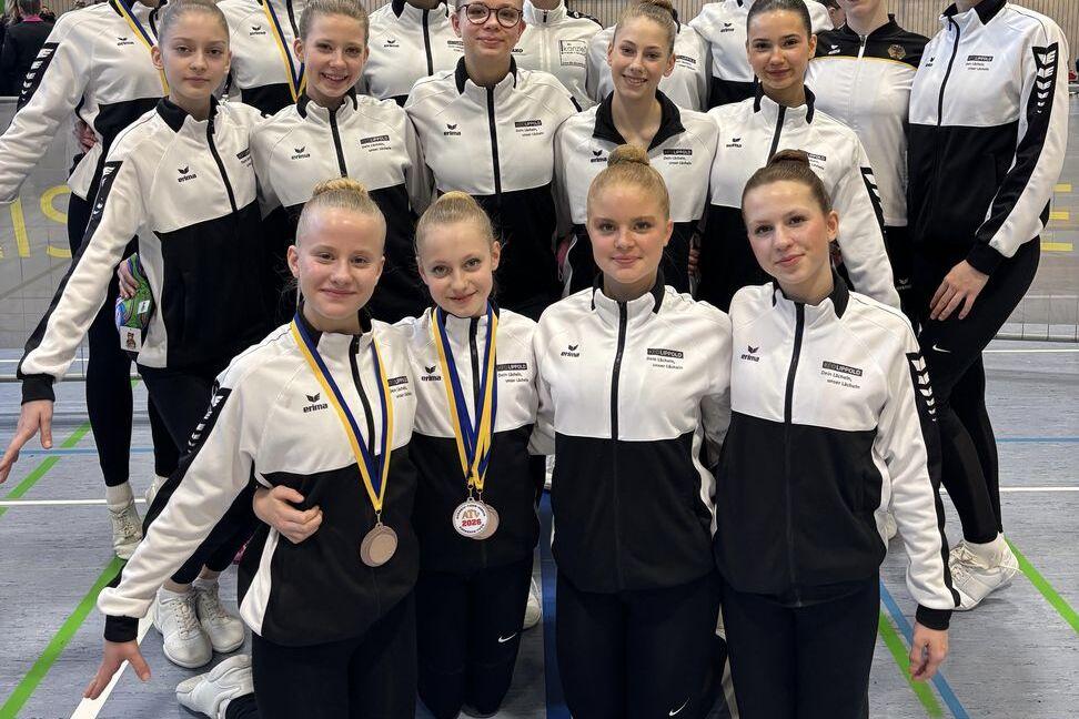 Gruppenshot von 15 Gymnastinnen in schwarzen und weißen Trainingsanzügen mit Medaillen, in einer Halle stehend und kniend.