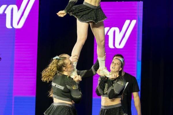 Cheerleaderin wird von zwei Teamkolleginnen in die Luft gehalten, während eine weitere Person im Hintergrund zuschaut.