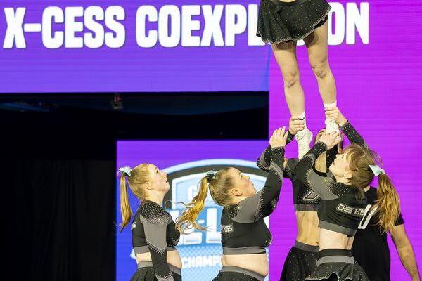 Fünf Cheerleader in glitzernden Outfits bilden einen Menschenstapel, während eine Athletin oben gehalten wird.
