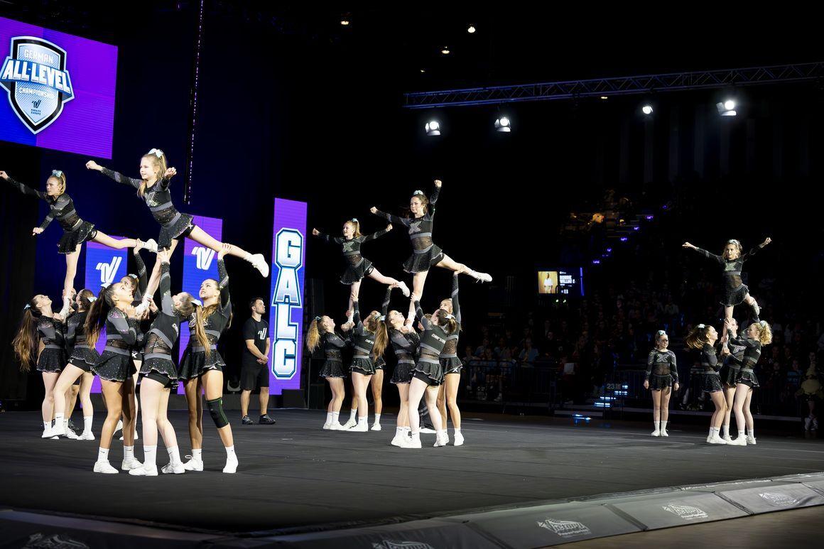Gruppenchoreografie mit Cheerleadern in schwarz, die Akrobatik und Stunts auf einer Bühne ausführen.