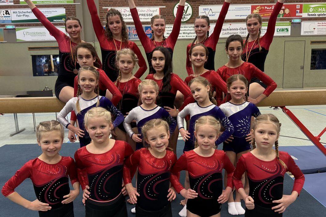 Gruppe von Turnerinnen in Gymnastikanzügen posiert auf einer Turnhalle, einige stehen hinter, andere sitzen vorne.