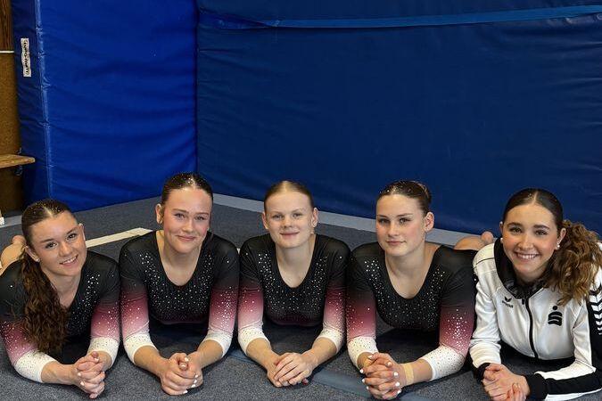 Fünf Mädchen in Sportoutfits liegen auf einer Gymnastikmatte in einer Turnhalle und posieren.