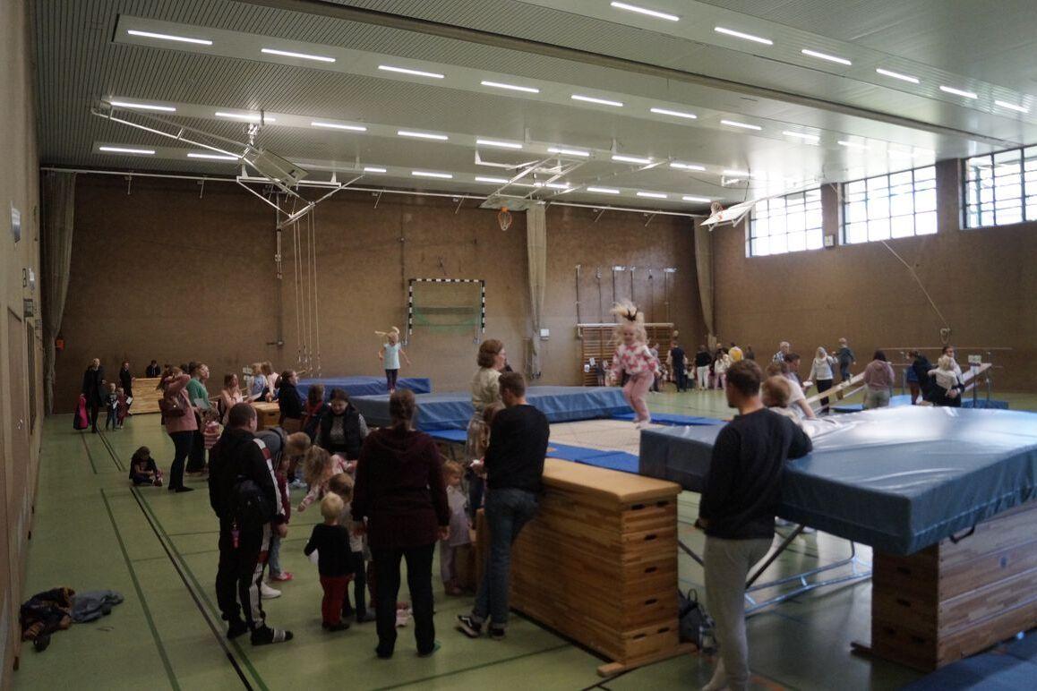 Gruppe von Kindern und Erwachsenen in einer Turnhalle, verschiedene Aktivitäten an Sprungtischen und Matten.