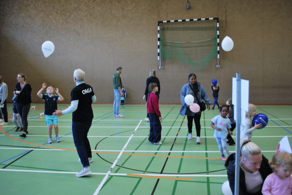 Gruppenspiele in einer Sporthalle mit Kindern, Erwachsenen und Luftballons auf einem grünen Sportboden.