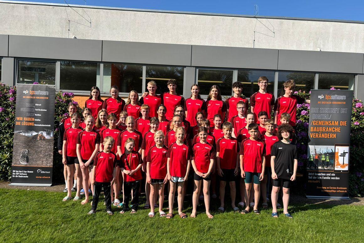 Gruppe von Sportlern in roten Shirts steht vor einem Gebäude mit Bannern, im Freien auf grünem Rasen.