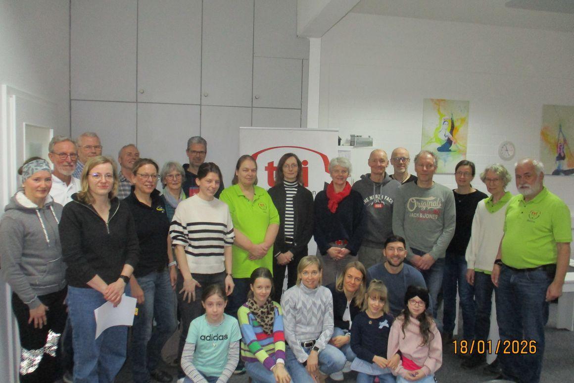 Gruppenfoto von 29 Personen, die in einem Raum posieren, einige tragen grüne Shirts, mit einem Banner im Hintergrund.