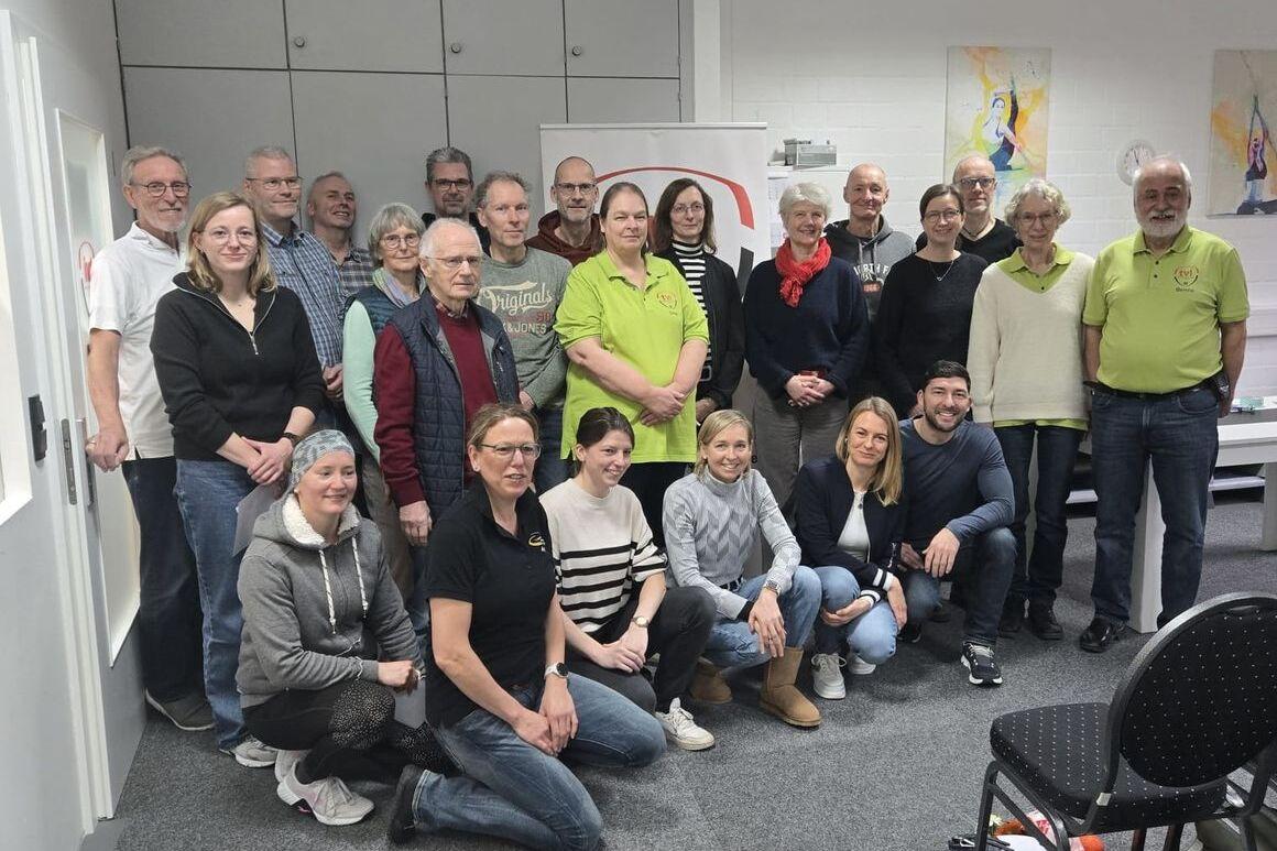 Gruppenfoto von 24 Personen in einem Raum, die lächeln, mit einem Banner im Hintergrund und Stühlen im Vordergrund.