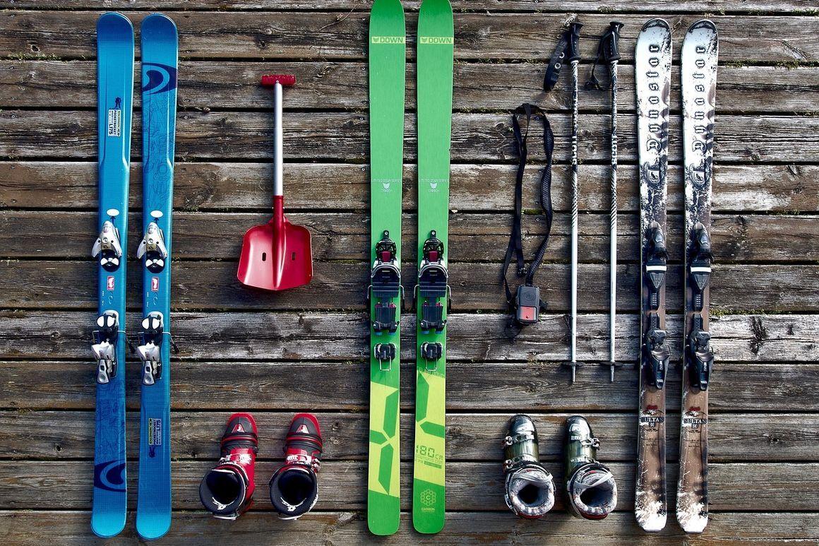 Ski-Equipment aus zwei Paar Skiern, Skischuhen, Skistöcken und einer Schaufel auf einem Holzuntergrund angeordnet.