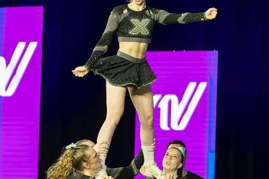 Cheerleaderin wird von zwei Teamkolleginnen in die Luft gehalten, w&auml;hrend eine weitere Person im Hintergrund zuschaut.