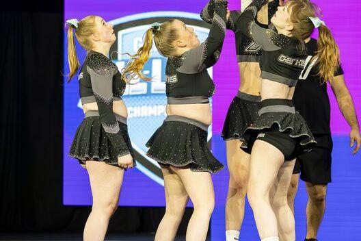 F&uuml;nf Cheerleader in glitzernden Outfits bilden einen Menschenstapel, w&auml;hrend eine Athletin oben gehalten wird.