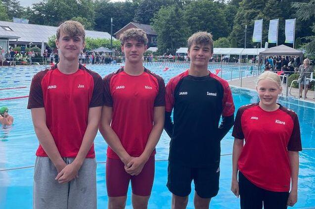 Vier junge Schwimmer in roten Sportoutfits stehen am Beckenrand eines Freibades, im Hintergrund sind Schwimmer und Zuschauer sichtbar.