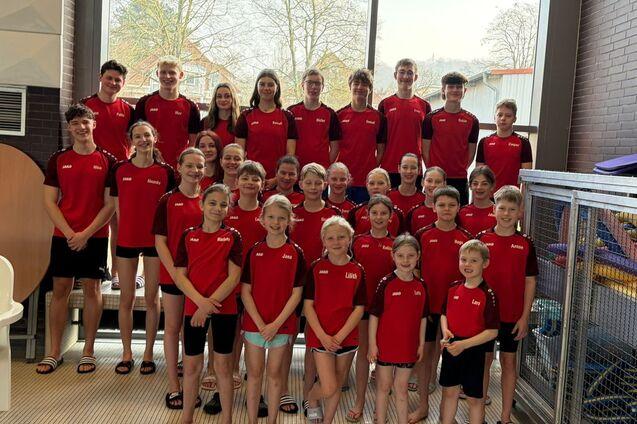 Gruppenfoto von Schwimmern in roten Trikots, aufgereiht vor großen Fenstern in einem Hallenbad.
