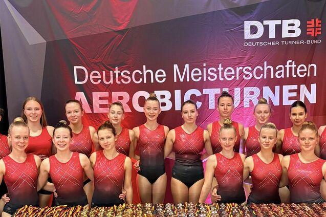 Gruppe von Sportlerinnen in roten Trikots vor einem Banner der Deutschen Meisterschaften im Aerobicturnen.