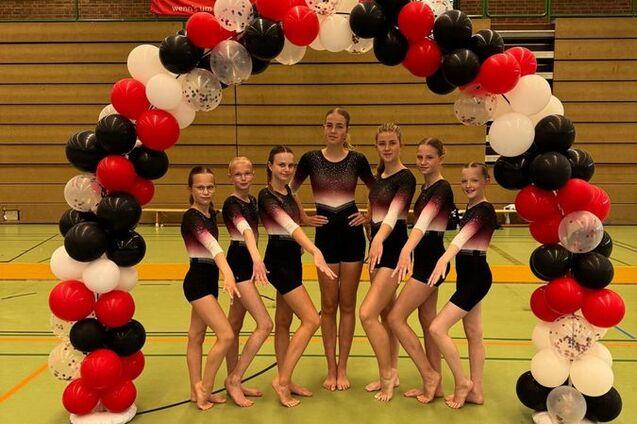 Gruppe junger Turnerinnen in Schwarz-Rot vor einem bunten Luftballonbogen in einer Sporthalle.