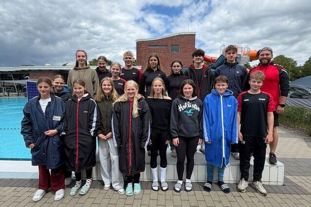 Gruppe von 20 Schwimmern in Umkleideanzügen steht vor einem Schwimmbecken, umgeben von einer Landschaft mit Wolken.