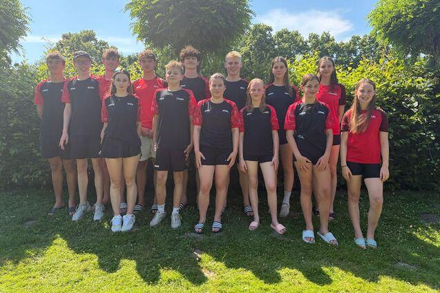 Gruppe von 15 Jugendlichen in roten und schwarzen Sportshirts, lächelnd im Freien vor einer grünen Hecke.