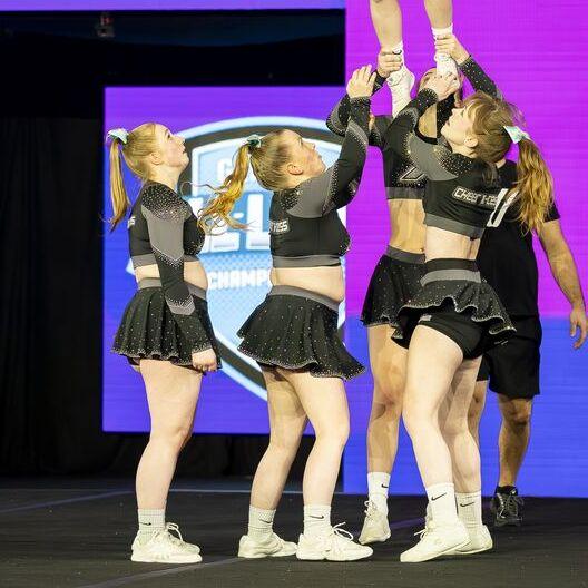 F&uuml;nf Cheerleader in glitzernden Outfits bilden einen Menschenstapel, w&auml;hrend eine Athletin oben gehalten wird.